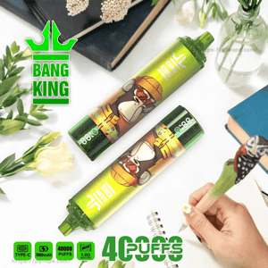 Two green Bang King disposable vape devices with a cartoon gorilla design lying on a white desk among books, flowers and a hand writing in a notebook, with 