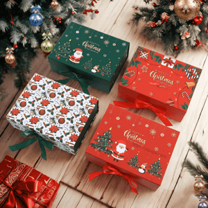 Four festive Christmas gift boxes—two red, one dark green with Santa and trees, and one white with poinsettia and ornament pattern—tied with red and green ribbons and arranged on a wooden floor beneath decorated evergreen branches with hanging baubles.